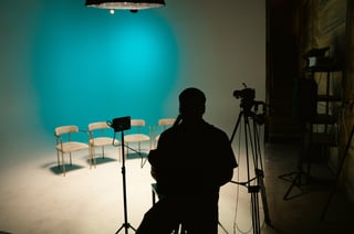 Silhouette of a person operating a camera on a film set with chairs, lights, and equipment against a turquoise backdrop