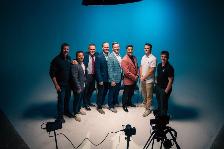 Group of eight men in business casual attire standing on a lit studio floor with professional lighting and camera equipment